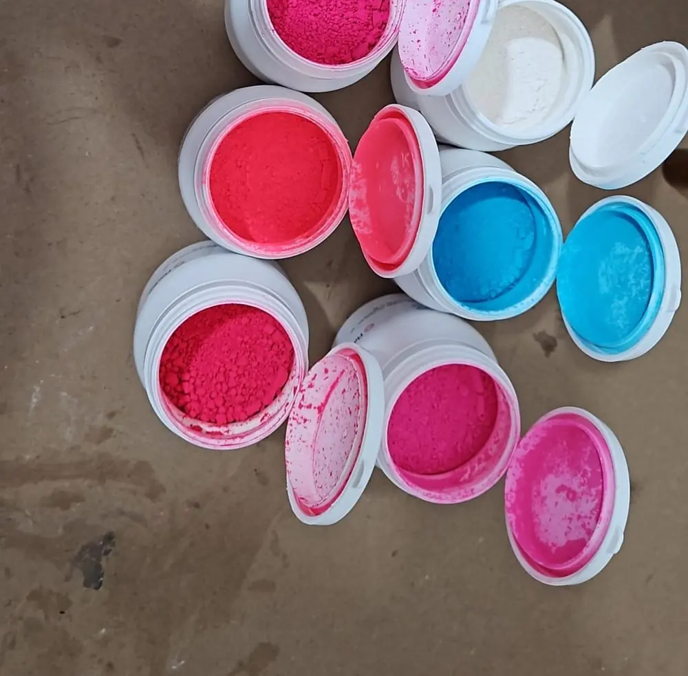 Barattoli aperti con pigmenti cosmetici in polvere di colore rosa, fucsia, arancio, azzurro e bianco, disposti su un piano di lavoro.